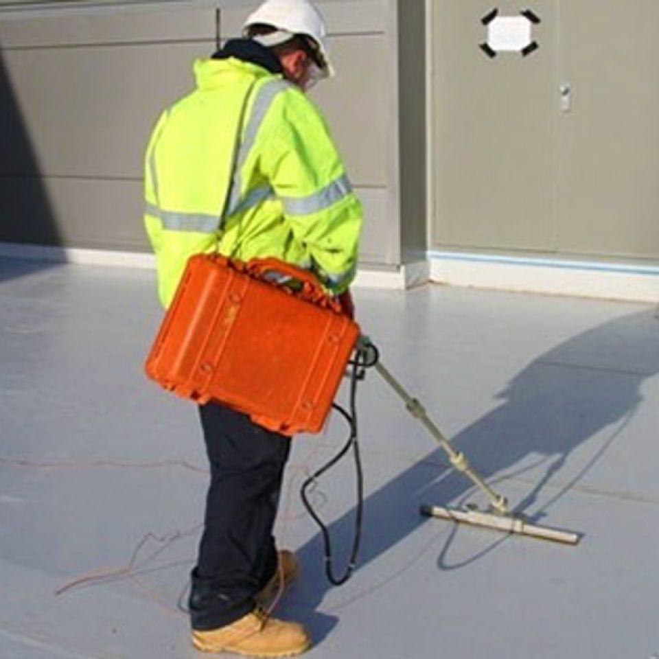 Technician in hi-vis carrying out a Dynamic Leak Investigation on a flat commercial roof using an electrical scanning tool.